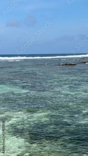 Weathered light gray wooden railing overlooks vibrant turquoise water with sandy bottom. Clear blue sky with scattered clouds completes tropical vista.