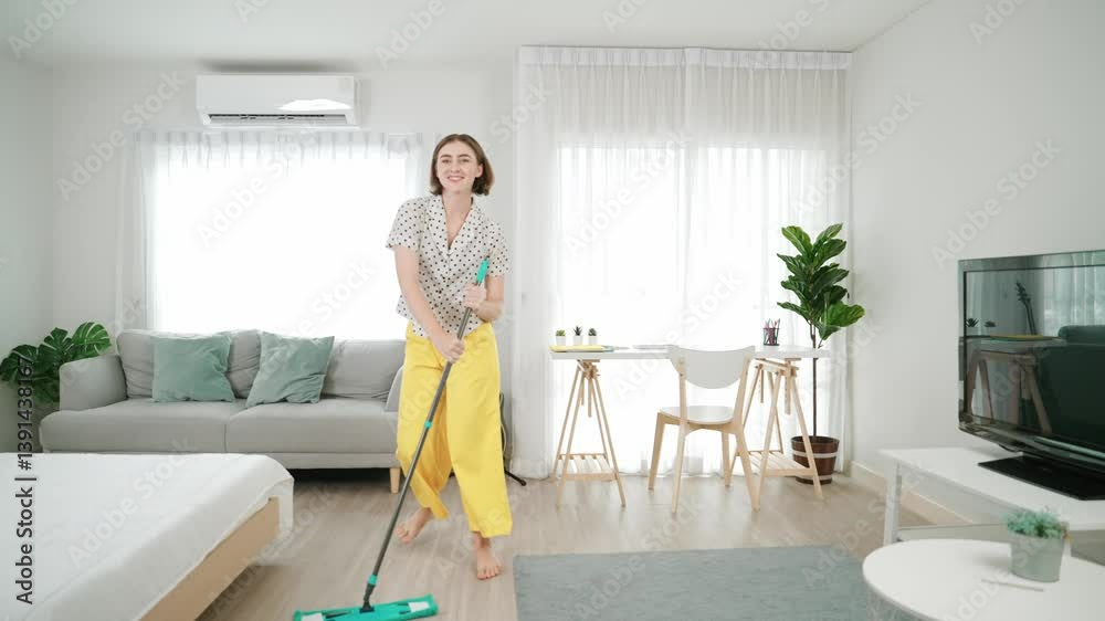 Caucasian housekeeper mopping the floor while dancing in lively mood at modern apartment. Happy energetic mother or cleaner enjoy cleaning the living room while moving to music. Lifestyle. Pedagogy. Caucasian housekeeper mopping the floor while dancing in lively mood at modern apartment. Happy energetic mother or cleaner enjoy cleaning the living room while moving to music. Lifestyle. Pedagogy.