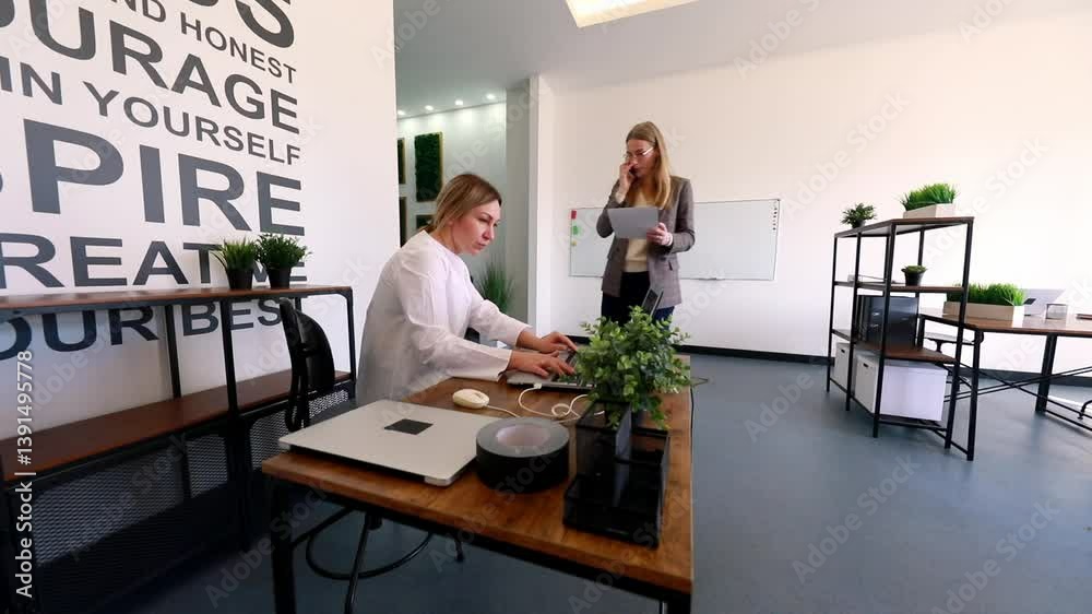 Women collaborating in a modern office space while one works on a laptop and the other reviews notes Women collaborating in a modern office space while one works on a laptop and the other reviews notes