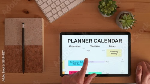 Businessman plans his weekly schedule, adding and moving appointments on a digital calendar using a touchscreen device, on a wooden desk with a keyboard, notepad, pencil, and plants