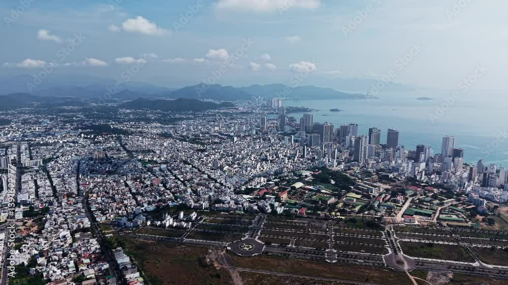 Aerial drone shot of the entire city of Nha Trang, Vietnam, with the ocean visible in the background. Captures a wide view of the urban landscape, coastline, and the bustling city life from above.