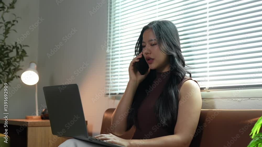 Frustrated young asian woman freelancer experiencing stress while talking angrily on phone and working intensively on laptop computer within home office environment Frustrated young asian woman freelancer experiencing stress while talking angrily on phone and working intensively on laptop computer within home office environment
