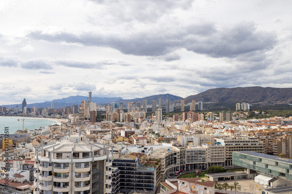 Aerial photo of the Spanish town of Benidorm in Spain showing the old town of the city with the beach known as Platja de Ponent or West Beach Promenade in background as well as mountains and clouds
