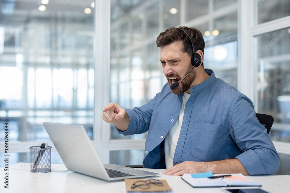 A furious customer service representative, wearing a headset, has an outburst in response to a problem on his laptop. He points and shouts with a displeased expression.