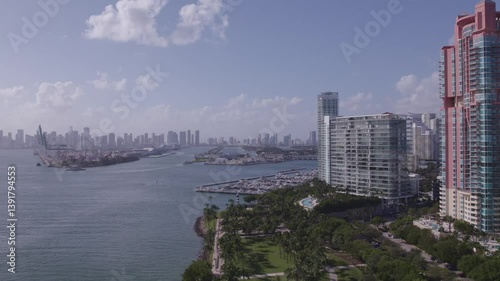 High aerial view over South Pointe Park with luxury towers, boats at marina, PortMiami cranes, and Downtown Miami skyline in the distance under sunny skies. photo