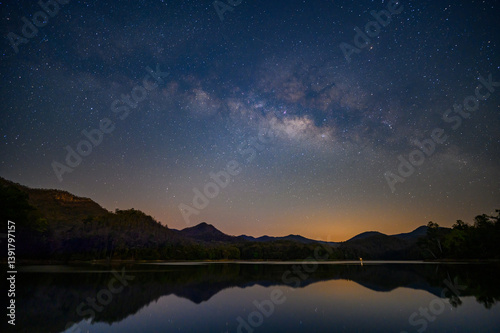 Fototapeta Naklejka Na Ścianę i Meble -  milky way stretches across the star-filled night sky above dark mountain silhouettes and a calm reflective lake, with a soft orange glow near the horizon