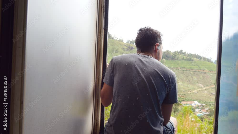 Unrecognizable guy with headphones sitting in entrance of moving train and leaning out from open doors. Young man riding on passenger railway transport and looking at beautiful nature. Rear back view Unrecognizable guy with headphones sitting in entrance of moving train and leaning out from open doors. Young man riding on passenger railway transport and looking at beautiful nature. Rear back view
