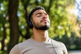 Close-up portrait of sportsman in park, hispanic man jogging in park with eyes closed breathing fresh air and resting, jogging with headphones listening to music and online radio and podcasts.