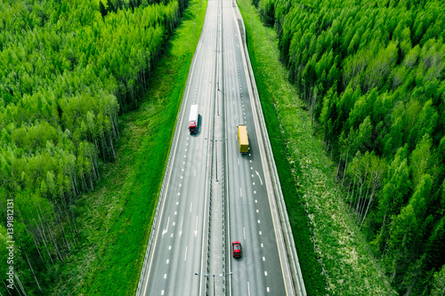 Obraz na płótnie Aerial view of highway with red car and modern semi trailers trucks, green trees and summer forests around w salonie