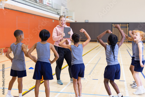 In school gym, female coach using tablet to instruct diverse children during fitness class