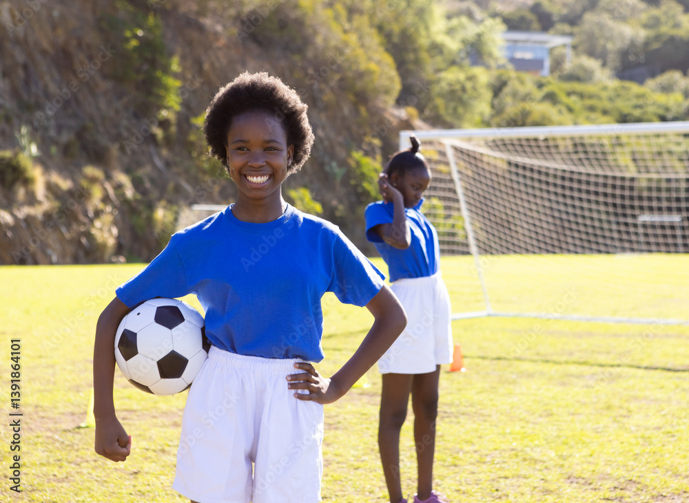 Obraz premium Smiling african american girl holding soccer ball on school field, teammate in background