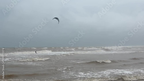 Kitesurfer riding waves on stormy sea under grey cloudy sky