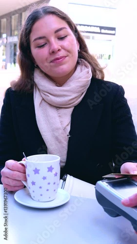young woman sits at an outdoor cafe, holding a coffee cup and paying with a credit card using a contactless payment terminal. She looks satisfied, representing modern finance and digital transactions photo