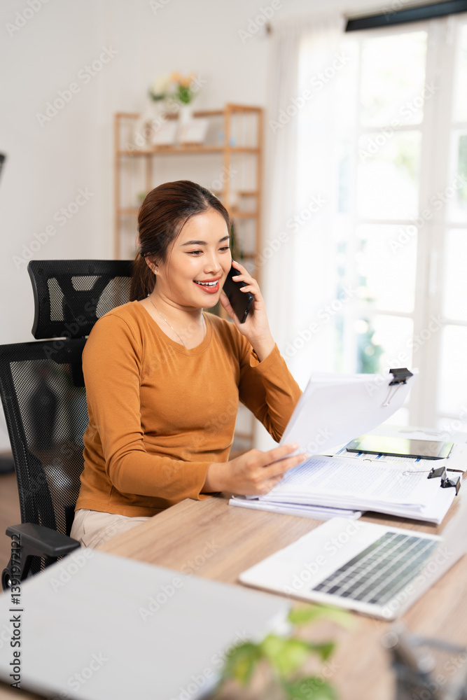 Young businesswoman cheerful asian lady in casual working in living room at home office, sitting by workdesk with laptop, holding documents, having phone conversation, copy space Young businesswoman cheerful asian lady in casual working in living room at home office, sitting by workdesk with laptop, holding documents, having phone conversation, copy space