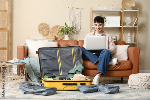 Male tourist using laptop on sofa while packing suitcase at home