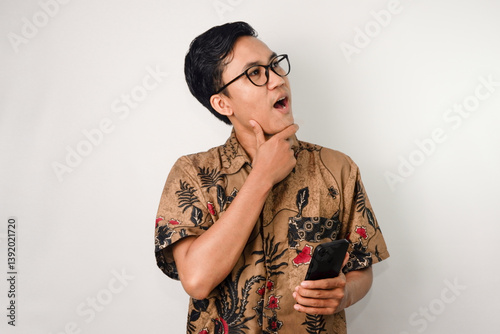 an asian man with glasses wearing a batik shirt holding a smartphone and posing as if having an aha moment