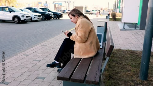 Young woman using smartphone while sitting on bench in urban area on bright sunny day