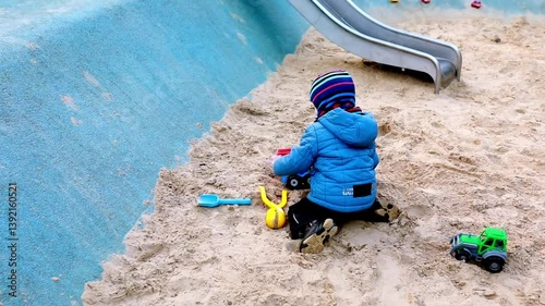 Child playing in sandbox with toy trucks near a playground slide on a sunny day in a local park