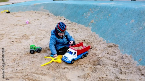 Child playing joyfully with toy trucks in a sandy park area during sunny weather