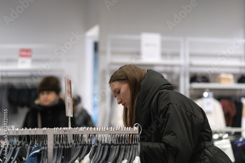 Young woman shopping for clothes, carefully examining garments on hangers in a clothing store, likely during a sale period, demonstrating mindful consumerism