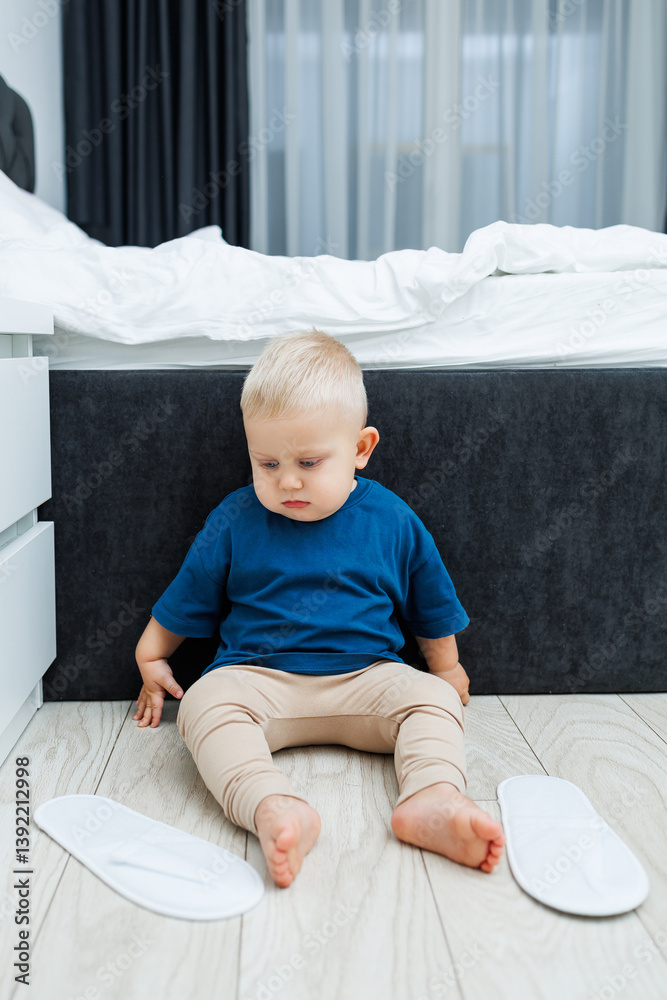 Cute blond toddler boy in blue t-shirt running in spacious hotel room. Toddler boy in comfortable hotel room resting after travel. Child at home sitting on floor