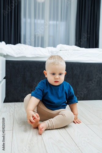 Cute blond toddler boy in blue t-shirt running in spacious hotel room. Toddler boy in comfortable hotel room resting after travel. Child at home sitting on floor
