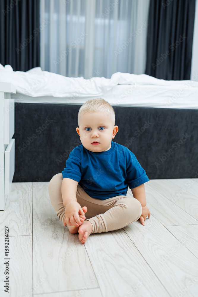 Cute blond toddler boy in blue t-shirt running in spacious hotel room. Toddler boy in comfortable hotel room resting after travel. Child at home sitting on floor