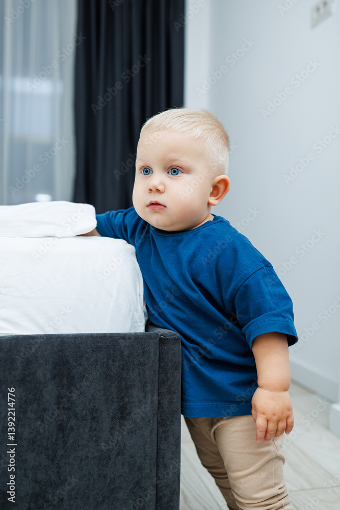 Cute blond toddler boy in blue t-shirt running in spacious hotel room. Toddler boy in comfortable hotel room resting after travel. Child at home sitting on floor
