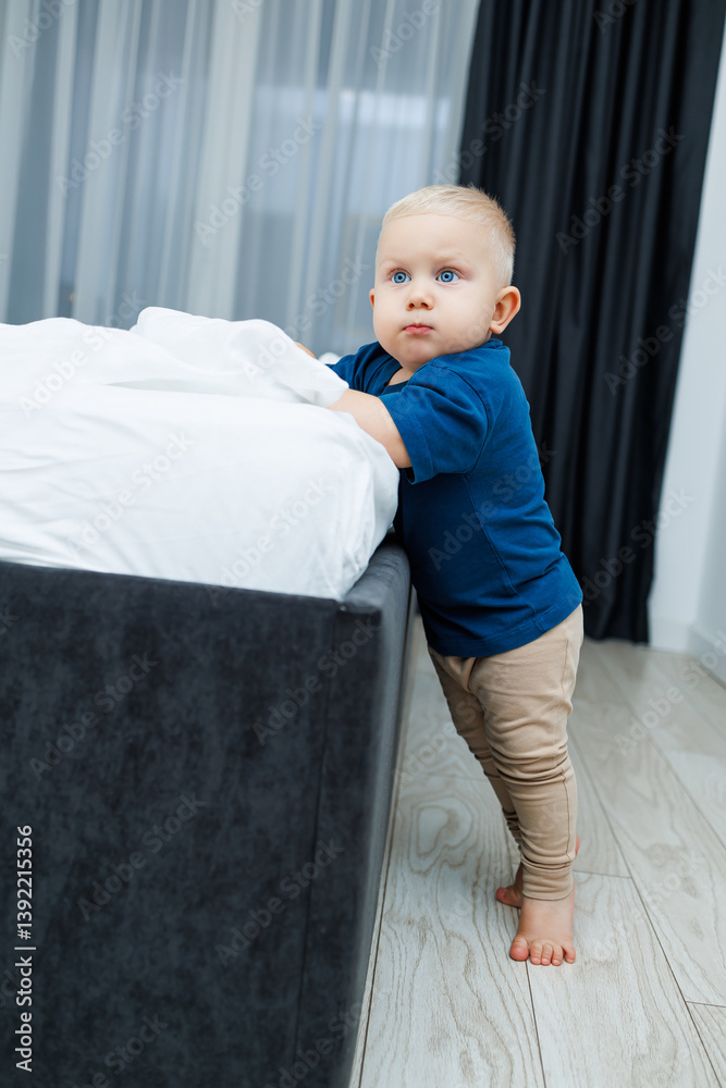 Cute blond toddler boy in blue t-shirt running in spacious hotel room. Toddler boy in comfortable hotel room resting after travel. Child at home sitting on floor