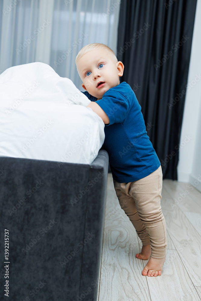 Cute blond toddler boy in blue t-shirt running in spacious hotel room. Toddler boy in comfortable hotel room resting after travel. Child at home sitting on floor