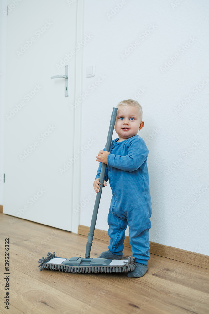 Toddler boy in blue romper cleans the room with a mop. Child washes the floor with a mop. Smiling boy runs around the house and cleans.