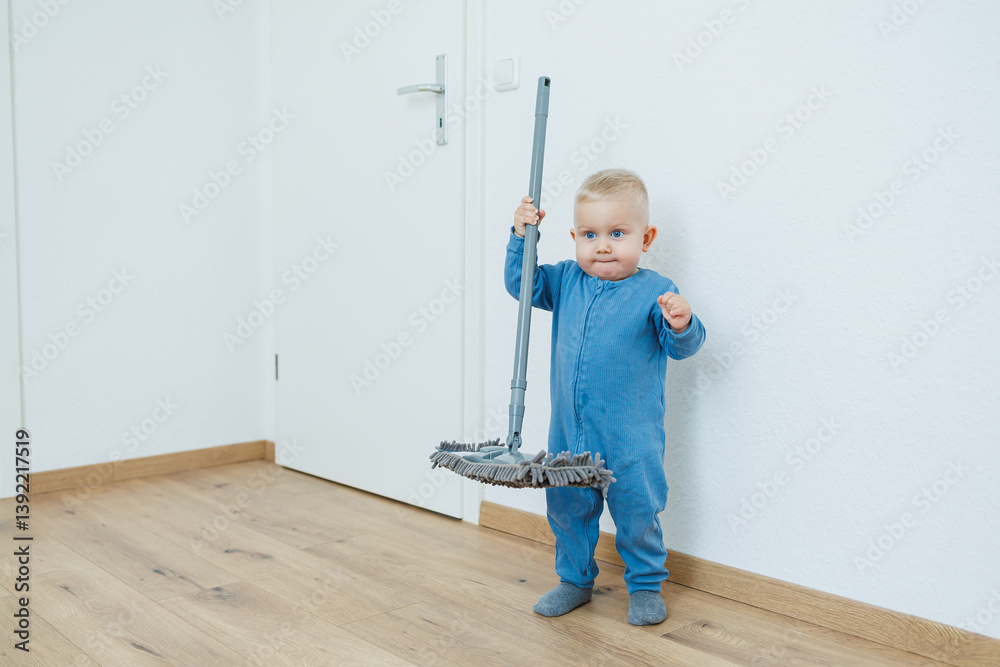 Toddler boy in blue romper cleans the room with a mop. Child washes the floor with a mop. Smiling boy runs around the house and cleans.