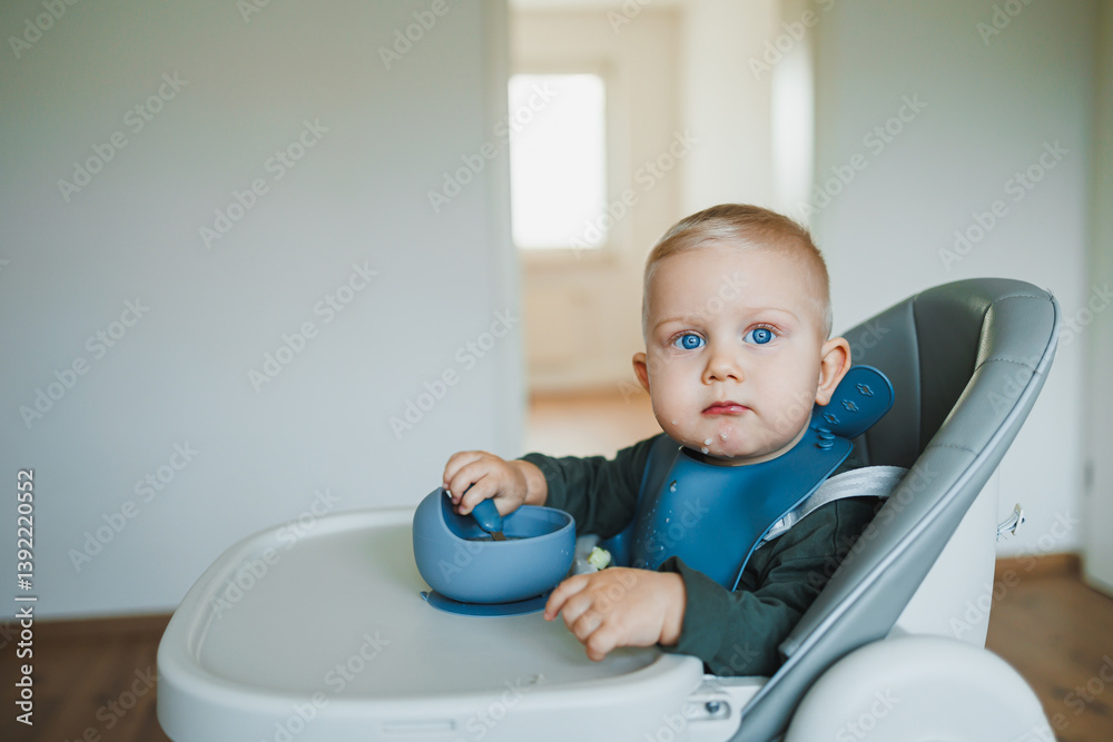 Smiling boy eats soup sitting on a chair. Hungry boy puts food in his mouth. Baby one year and three months old eats independently. Silicone utensils for feeding children.