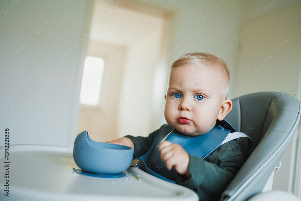 A one and a half year old boy sits on a high chair and independently eats soup from a silicone plate with a spoon. Silicone utensils for feeding children. The child learns to eat independently.