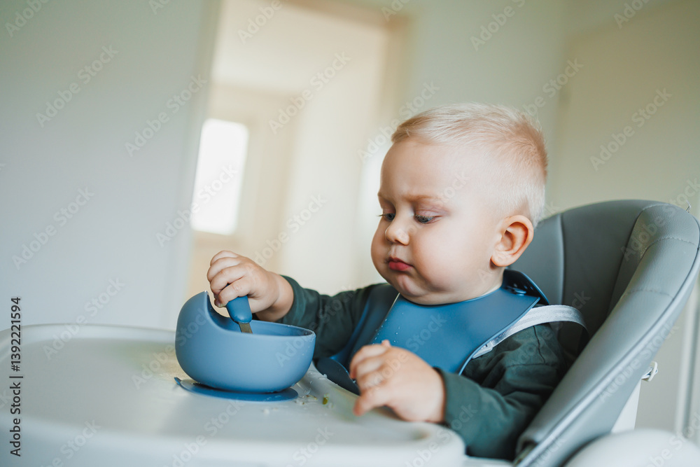 Smiling boy eats soup sitting on a chair. Hungry boy puts food in his mouth. Baby one year and three months old eats independently. Silicone utensils for feeding children.