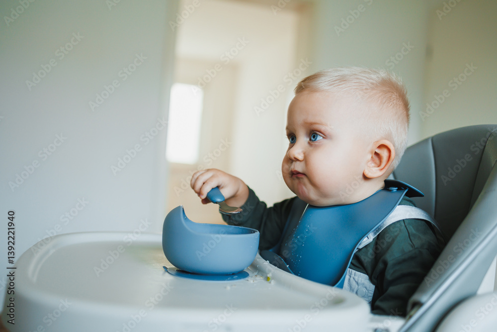 A one and a half year old boy sits on a high chair and independently eats soup from a silicone plate with a spoon. Silicone utensils for feeding children. The child learns to eat independently.