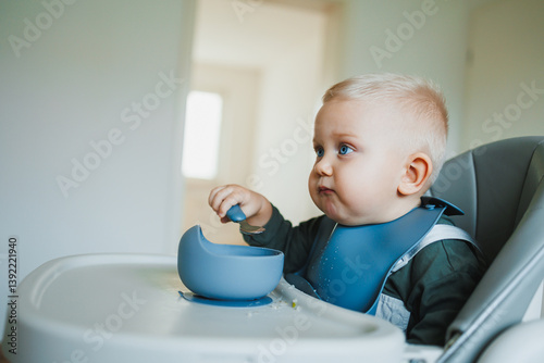 Obraz na płótnie A one and a half year old boy sits on a high chair and independently eats soup from a silicone plate with a spoon. Silicone utensils for feeding children. The child learns to eat independently. w salonie