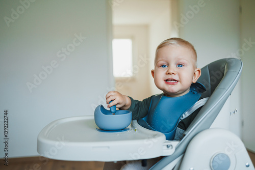 Smiling boy eats soup sitting on a chair. Hungry boy puts food in his mouth. Baby one year and three months old eats independently. Silicone utensils for feeding children.