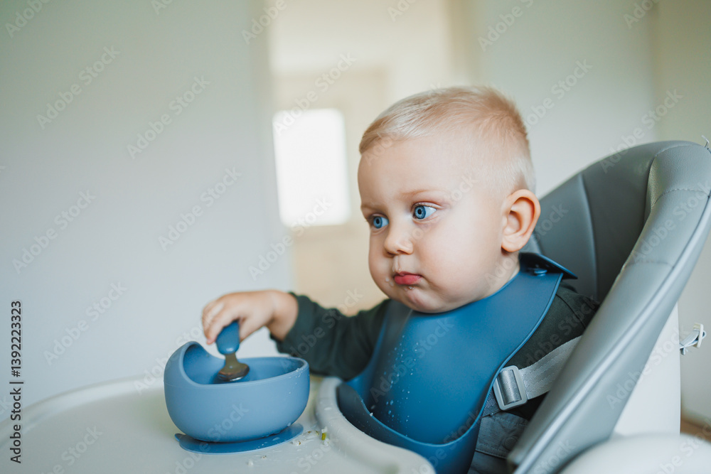 Smiling boy eats soup sitting on a chair. Hungry boy puts food in his mouth. Baby one year and three months old eats independently. Silicone utensils for feeding children.