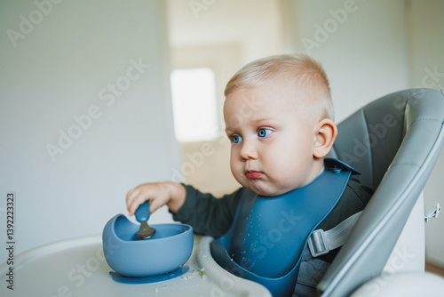 Obraz na płótnie Smiling boy eats soup sitting on a chair. Hungry boy puts food in his mouth. Baby one year and three months old eats independently. Silicone utensils for feeding children. w salonie