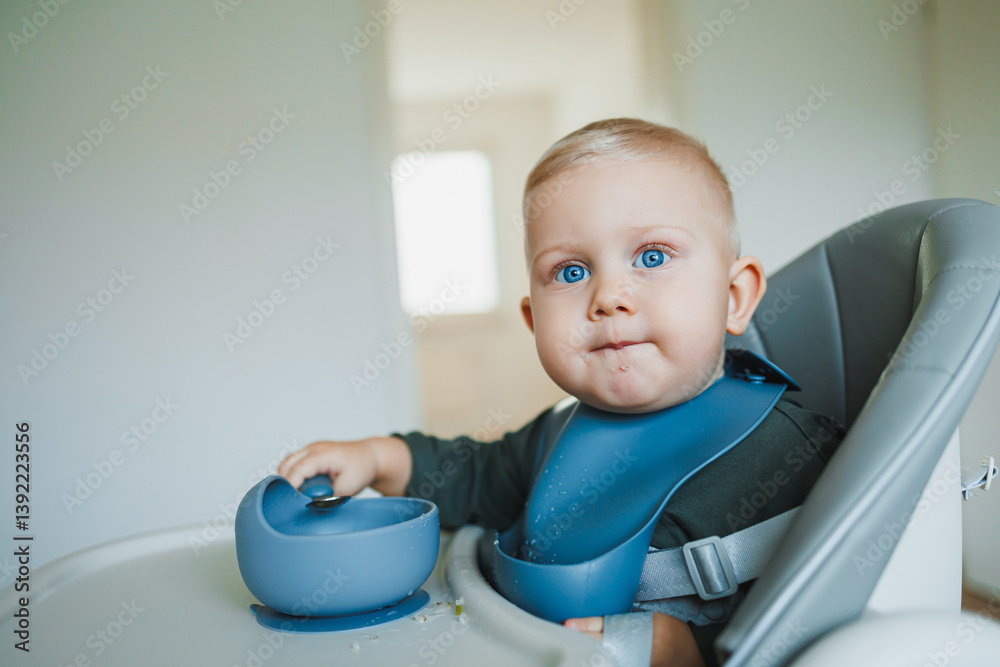 Toddler boy in casual clothes sits at the table in a highchair and eats himself with a spoon. The child learns to eat independently. Healthy nutrition for children. Baby food