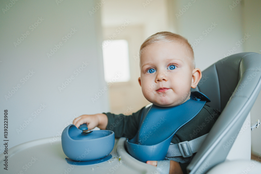 Blue-eyed toddler boy sitting on a chair with a silicone apron eating independently. Toddler boy learning to eat independently. Safe utensils for feeding children.