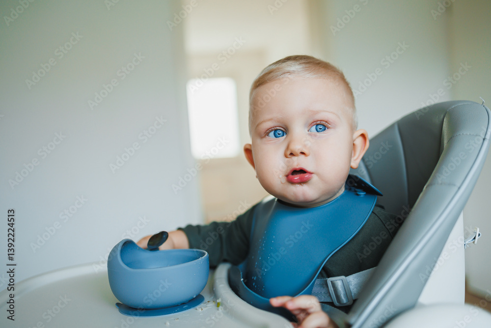 Blue-eyed toddler boy sitting on a chair with a silicone apron eating independently. Toddler boy learning to eat independently. Safe utensils for feeding children.