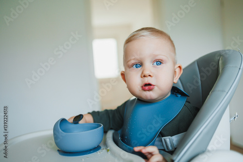 Obraz na płótnie Blue-eyed toddler boy sitting on a chair with a silicone apron eating independently. Toddler boy learning to eat independently. Safe utensils for feeding children. w salonie