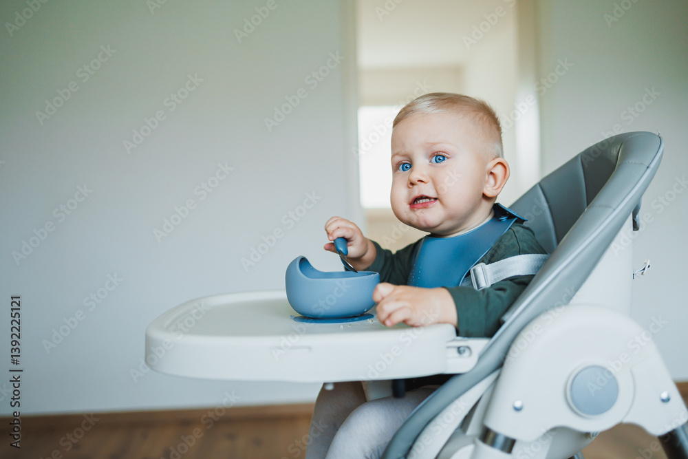 Blue-eyed toddler boy sitting on a chair with a silicone apron eating independently. Toddler boy learning to eat independently. Safe utensils for feeding children.