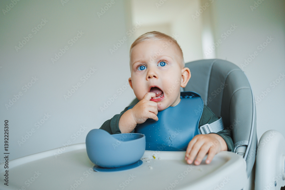 Blue-eyed toddler boy sitting on a chair with a silicone apron eating independently. Toddler boy learning to eat independently. Safe utensils for feeding children.