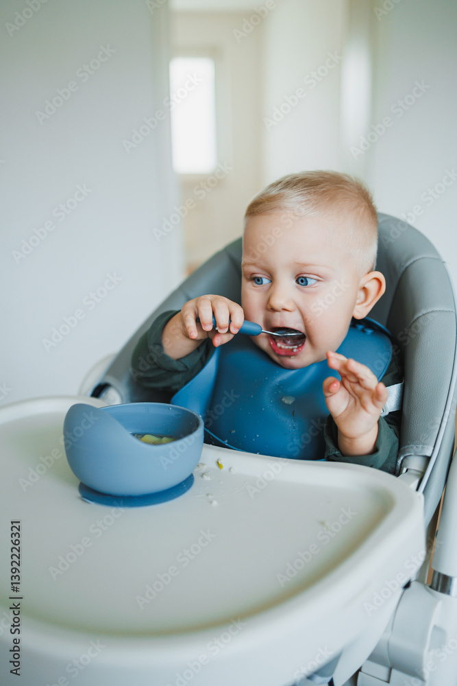 Toddler boy in casual clothes sits at the table in a highchair and eats himself with a spoon. The child learns to eat independently. Healthy nutrition for children. Baby food