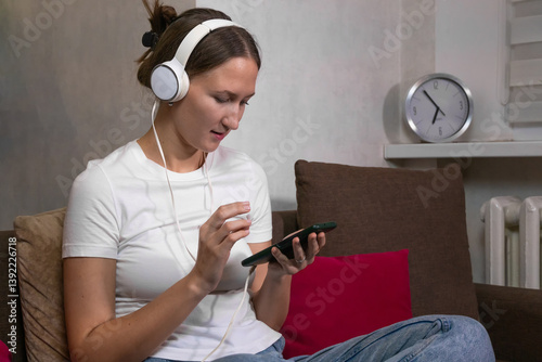 Obraz na płótnie Young woman wearing white headphones is listening to music and browsing her smartphone while sitting on a comfortable sofa in her living room w salonie