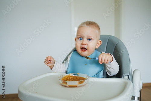 Blue-eyed toddler boy sitting on a chair with a silicone apron eating independently. Toddler boy learning to eat independently. Safe utensils for feeding children.