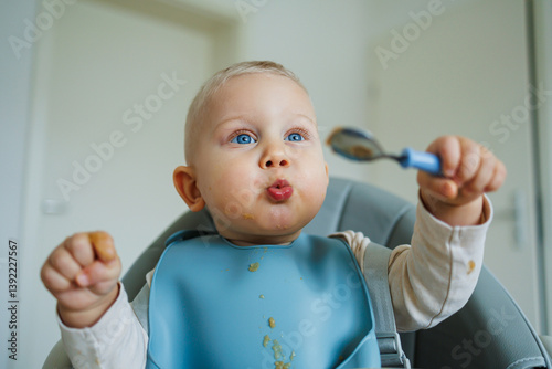 Blue-eyed toddler boy sitting on a chair with a silicone apron eating independently. Toddler boy learning to eat independently. Safe utensils for feeding children.